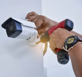 A person installs a security camera on a white wall using a red cordless drill. The persons hands and watch are visible, focusing on attaching the camera bracket.