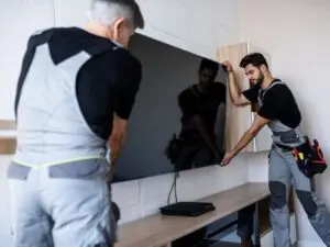Two men in work uniforms carefully mount a flat-screen TV on a wall above a wooden console. One man steadies the TV from the front, while the other supports it from the side, demonstrating general handyman services offered by BrightWise Handyman Services.