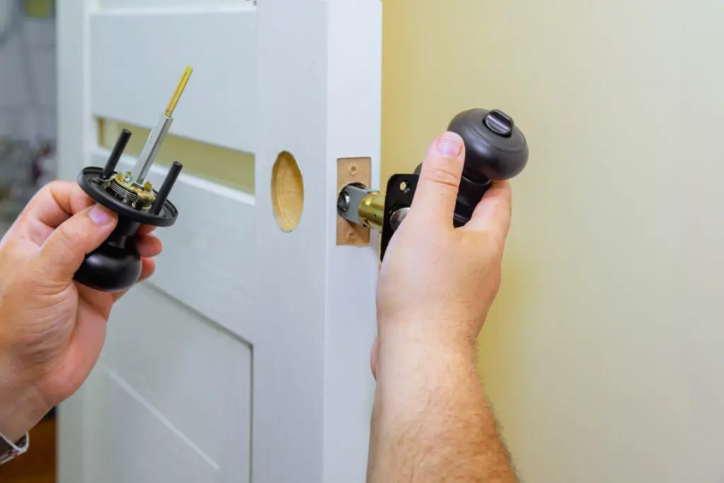 A person installs a black door handle and lock on a white door, holding parts of the doorknob set on each side of the door near the latch hole.
