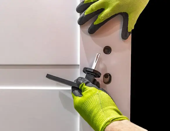 A person wearing bright green gloves installs a door handle and lock, holding the handle with one hand and using a tool with the other hand against a white door.