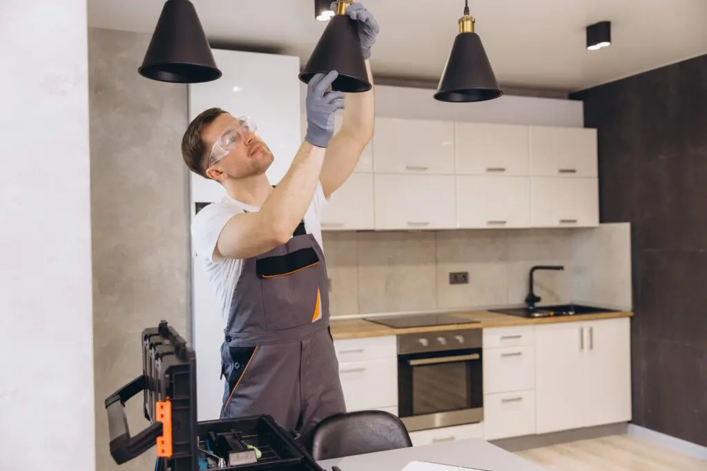 A man wearing safety goggles and gloves installs or fixes a pendant light fixture in a modern kitchen. A toolbox is on the counter nearby.
