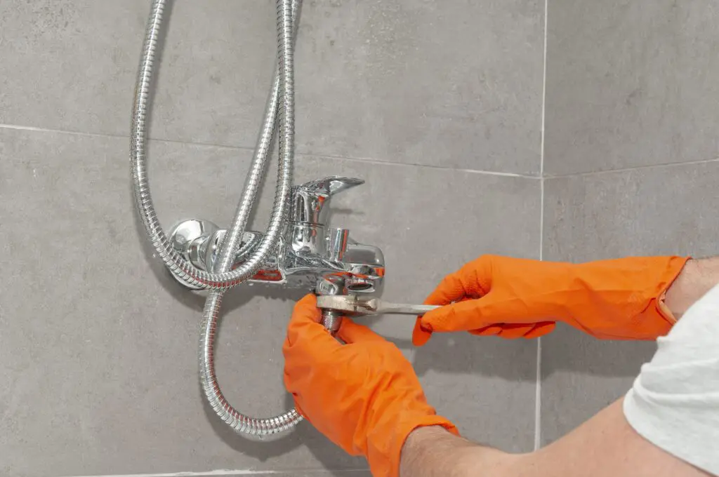 A person wearing orange gloves uses a wrench to tighten or repair a chrome shower faucet attached to a tiled bathroom wall.