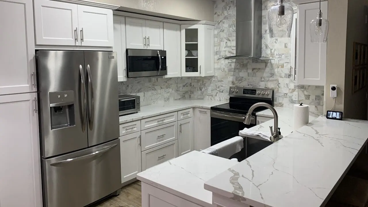 Modern kitchen with white cabinets, stainless steel appliances, marble backsplash and countertops, a sink with a towel, and pendant lighting above the island. Refrigerator, microwave, and oven are visible.