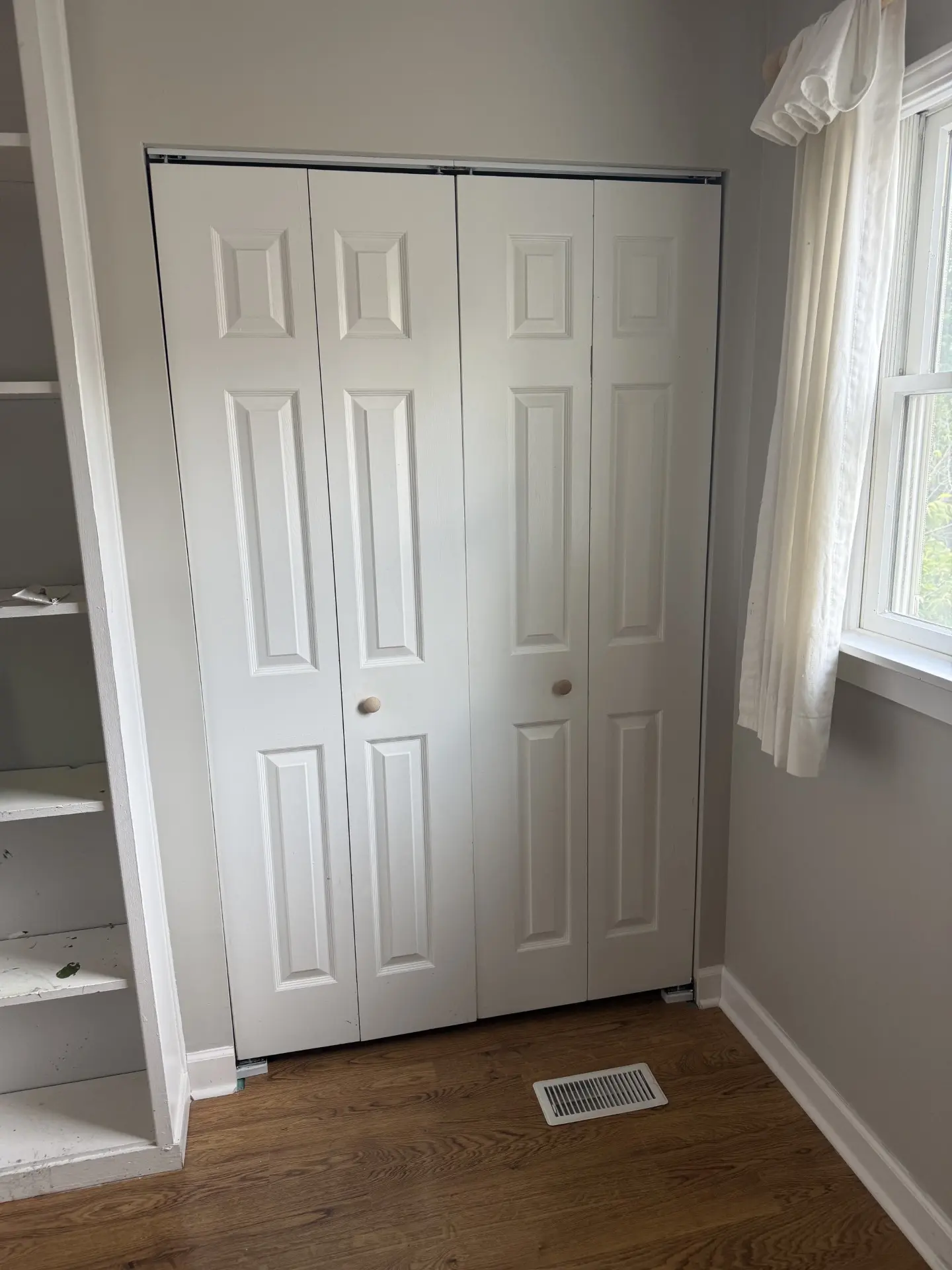 Naperville-closet-doors A pair of white bi-fold closet doors with wooden knobs, next to a window with white curtains and a built-in white shelf. The room has light gray walls and a wooden floor with a vent.