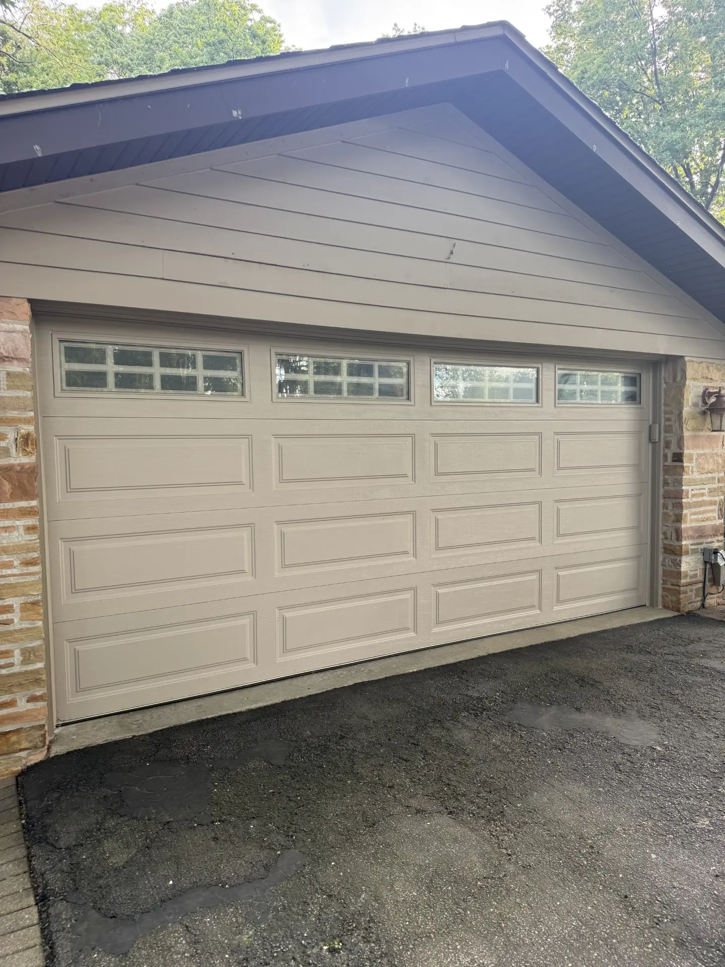 Garage Door Molding Replacement in Naperville Il A closed beige garage door with four rectangular windows at the top, set in a brick and siding exterior, with an asphalt driveway in front. Trees and cloudy sky are visible in the background.
