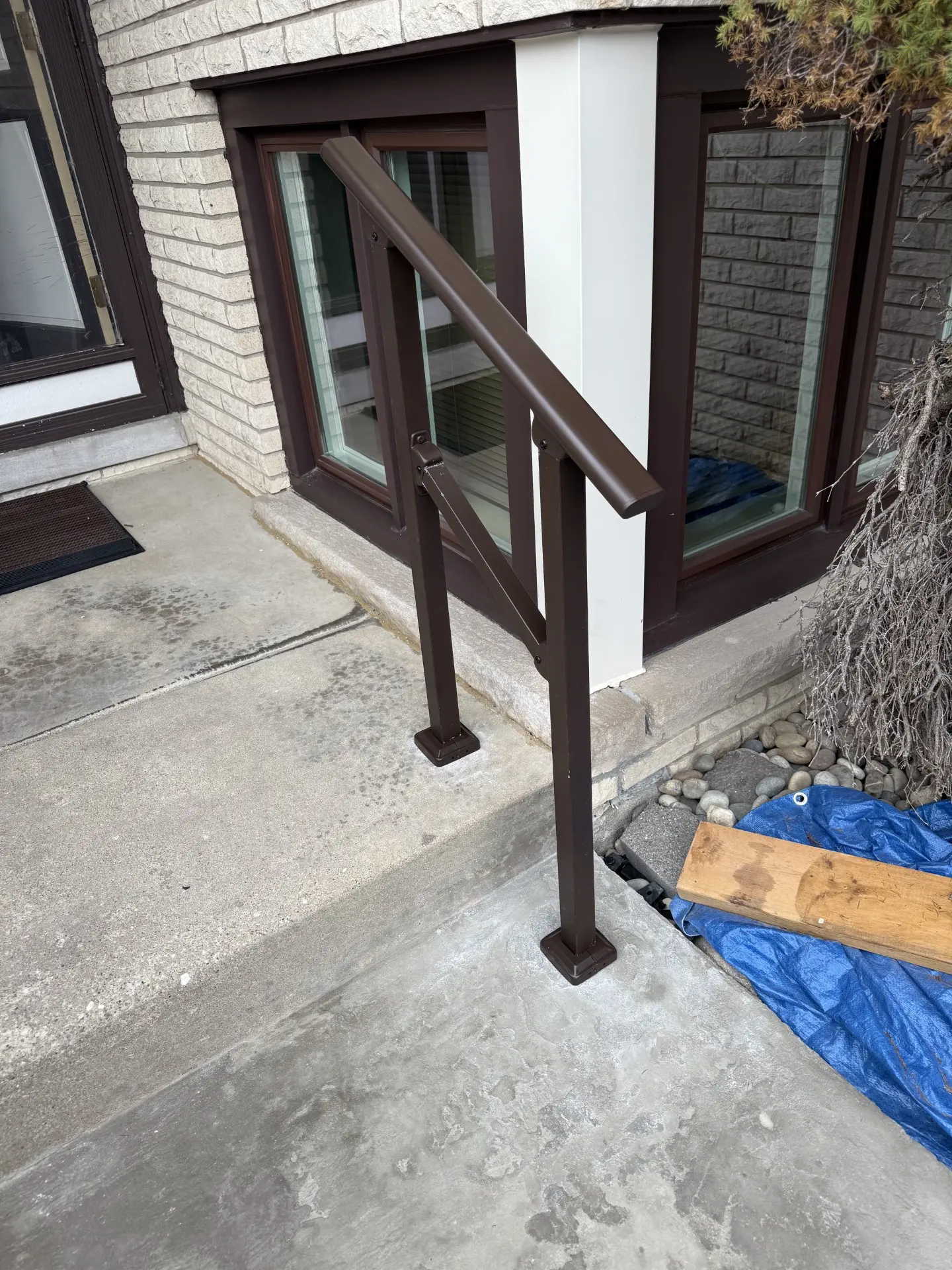 A brown metal handrail is installed alongside a concrete step, next to a window-well with white trim. A blue tarp, rocks, and a wooden plank are on the right side of the image near the house.