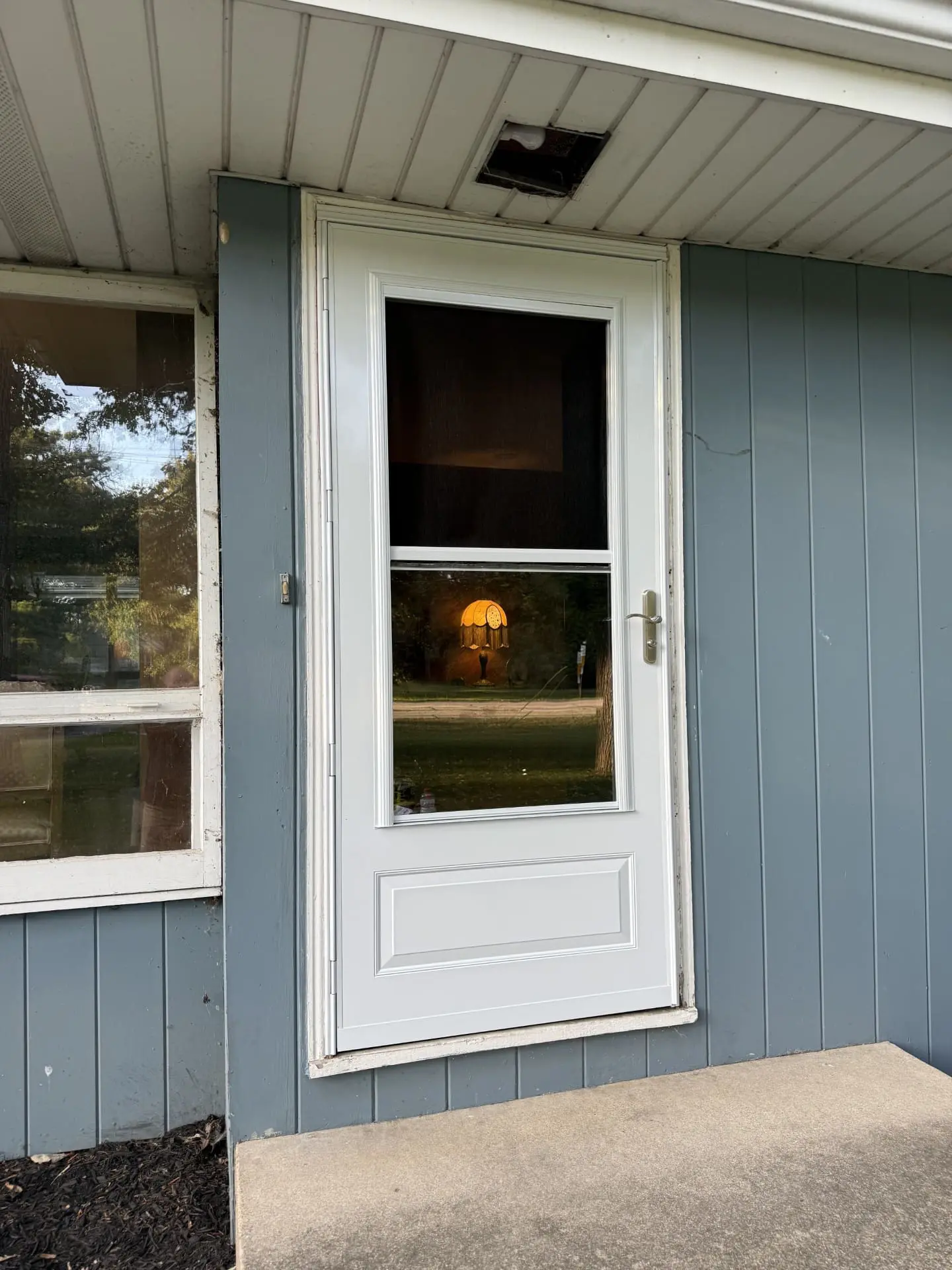 A white screen door on a blue-gray house with a concrete step in front; the doors glass reflects trees and a lamp is visible through an interior window. A damaged light fixture is above the door.
