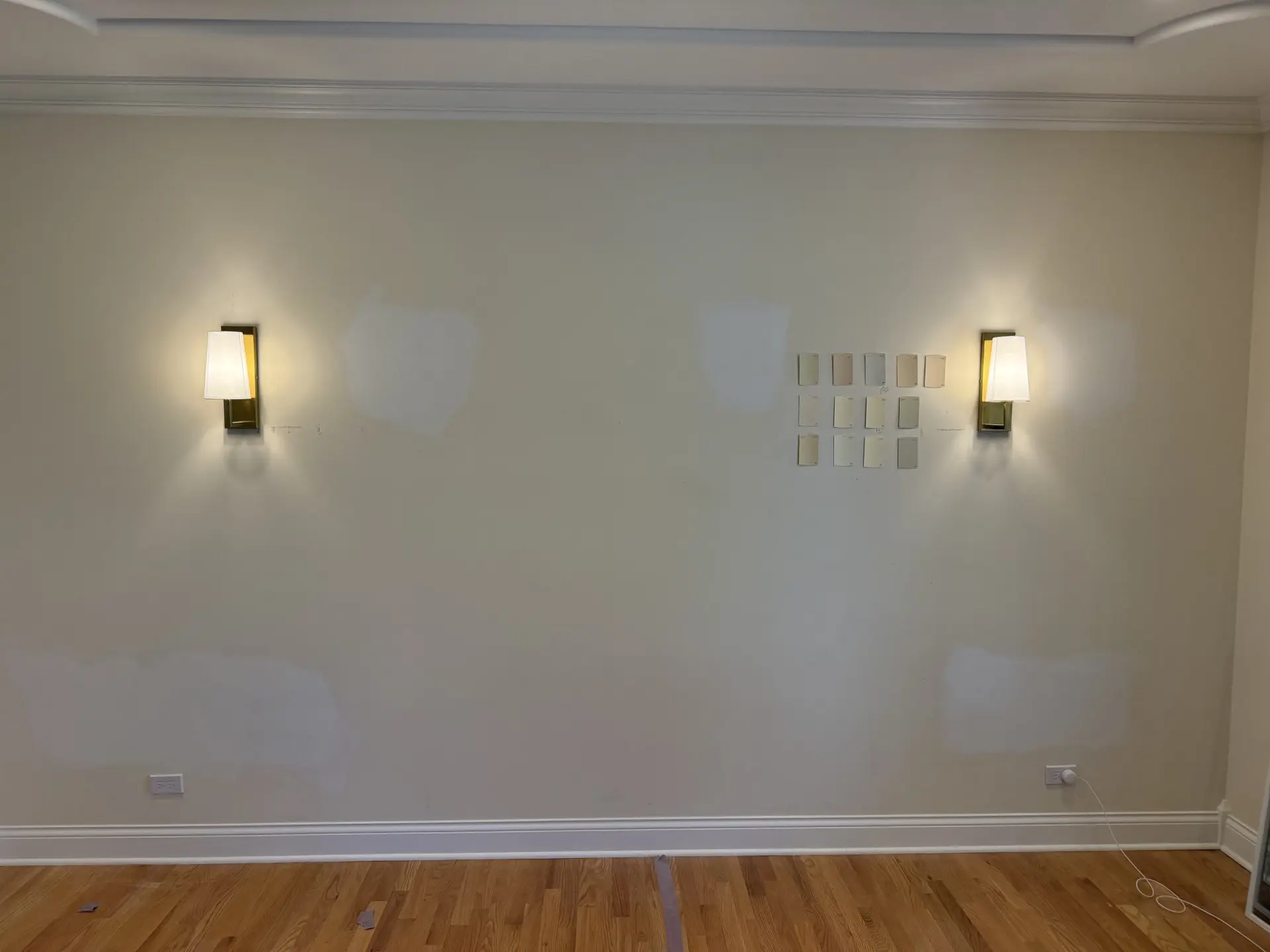 A beige wall with two sconces and several white paint swatches in the center. There are visible patches of spackling on the wall, light wood flooring, and white crown molding along the top and bottom edges.
