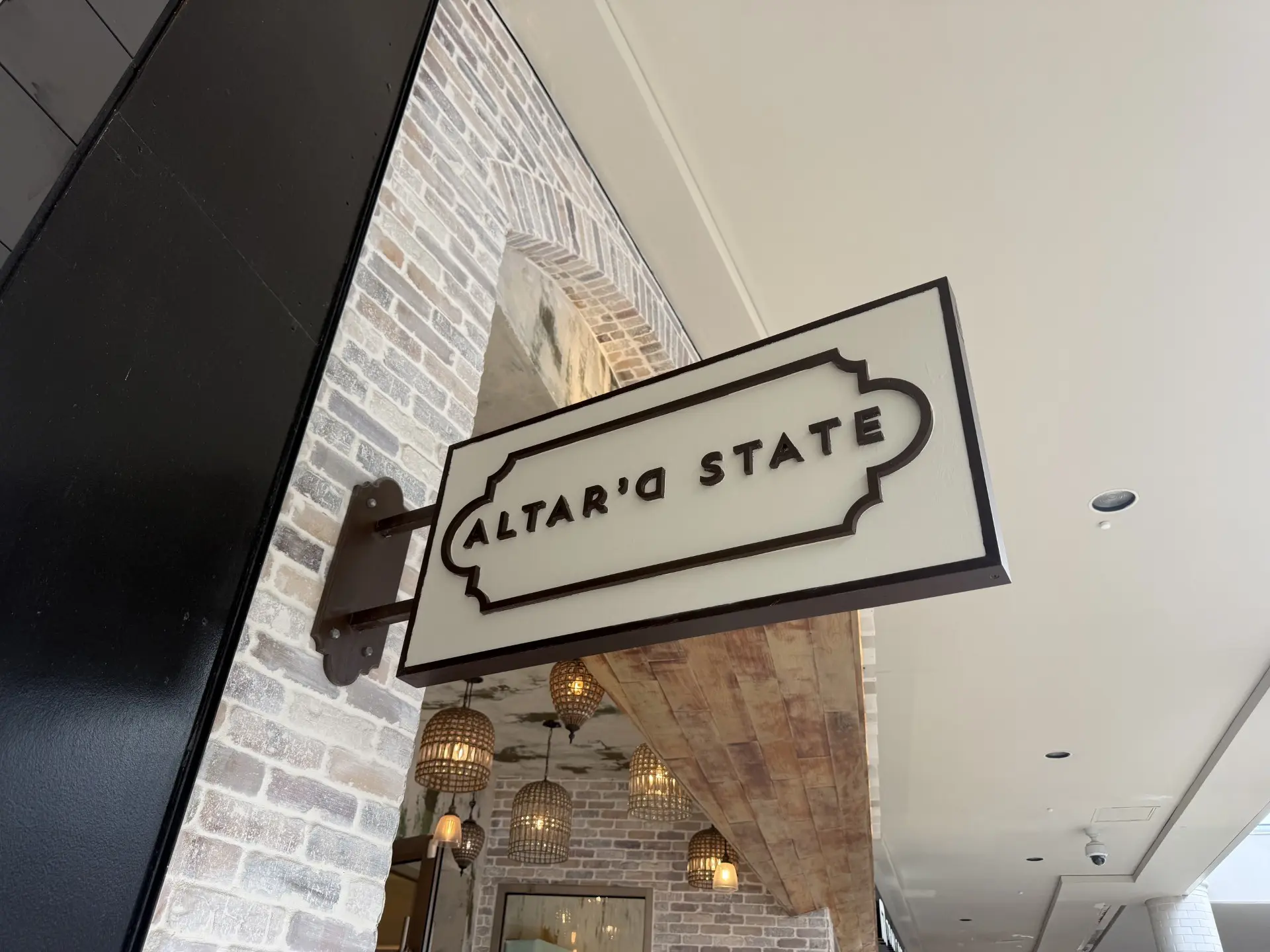 Alter'd State Retail Store Sign in Oak Brook Center A retail store sign reads ALTARD STATE in bold letters, mounted on a brick wall with arched details. Decorative hanging lights and ceiling reflect in the background.