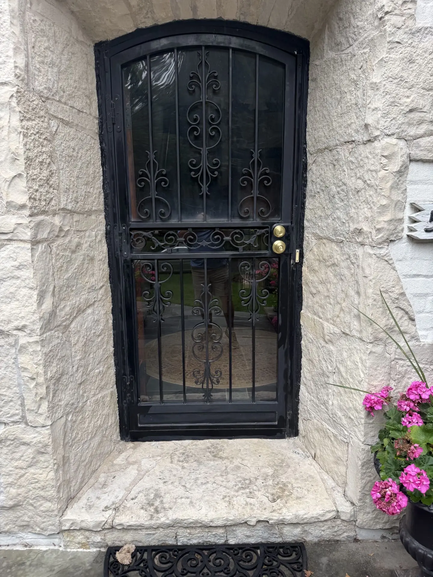 A black wrought iron door with decorative scrollwork set in a stone entryway. Pink flowers in a pot sit to the right of the doorway on a step, and part of a doormat is visible at the bottom.