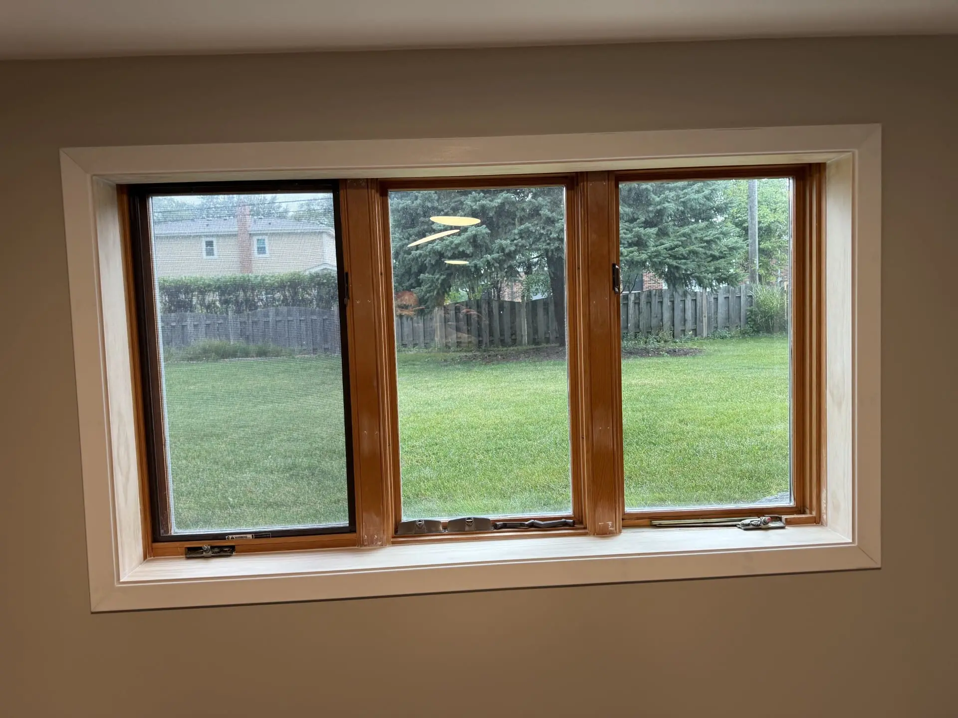 A wooden-framed window with three glass panes looks out onto a backyard with green grass, a wooden fence, and trees. The sky is overcast, and there are houses visible beyond the fence.