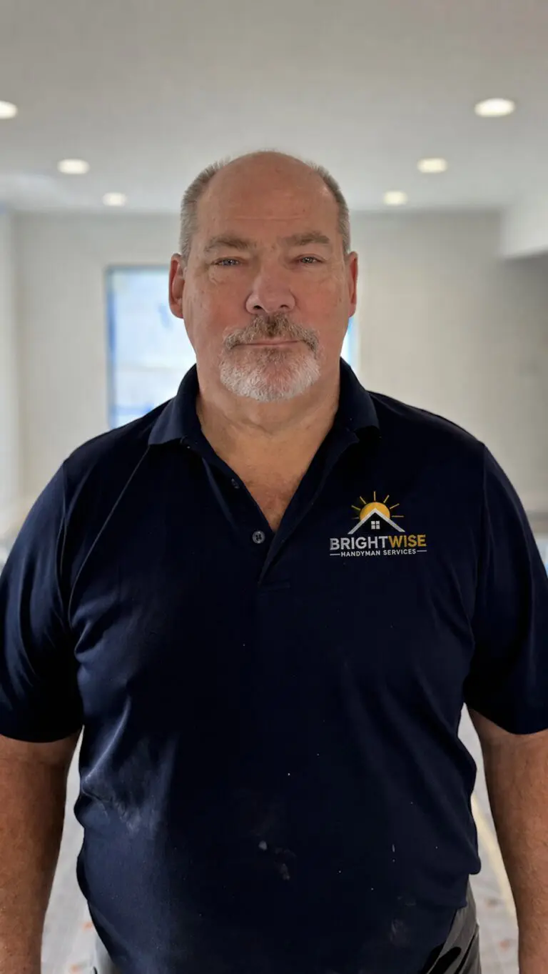 A man with a mustache and beard stands indoors, wearing a navy blue Brightwise Handyman Service polo shirt. The background is out of focus with soft lighting and light-colored walls.