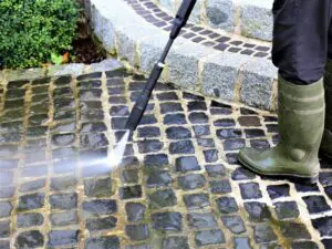 A person in green boots uses a pressure washer to clean a cobblestone path near stone steps and greenery.