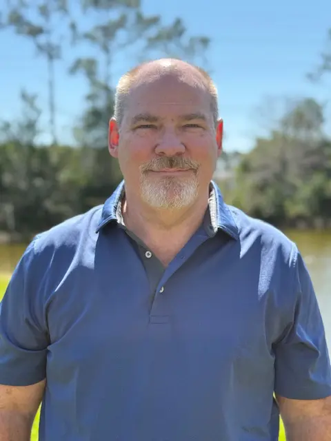 A man with short hair, a beard, and mustache stands outdoors in front of a pond and trees, wearing a blue collared shirt and smiling at the camera on a sunny day.