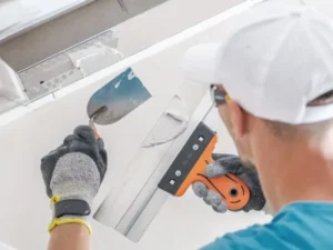A person wearing gloves and a cap uses a trowel and a putty knife to apply plaster to a wall, expertly working on a drywall joint as part of professional drywall services.