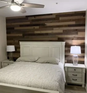 A bedroom with a white bed and matching nightstands, each with a clear glass lamp. The accent wall features horizontal wooden planks in varying browns. A ceiling fan is above—perfect results you’d expect from Brightwise Handyman services near you.