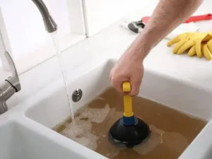 A person uses a plunger to unclog a kitchen sink filled with dirty water, with rubber gloves, a wrench, and plumbing services pamphlets on the countertop nearby.