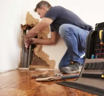 A person kneels on a wooden floor repairing exposed pipes in a wall, with torn drywall around the area; tools and a tool bag from Brightwise Handyman are in the foreground.