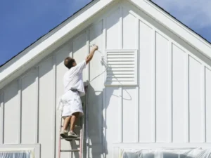 A person stands on a ladder, wearing white clothing, and uses a paint sprayer to provide painting services on the exterior wall of a white house under a blue sky. The windows are covered with protective plastic sheets.