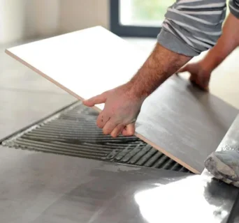 A handyman is laying a large ceramic tile onto a floor, spreading adhesive underneath. The persons hands are visible as they carefully position the tile into place.
