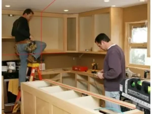 Two men working in a kitchen under renovation; one stands on a ladder near cabinets, while the other checks a device. A laser level projects red lines across the room for alignment during carpentry services.
