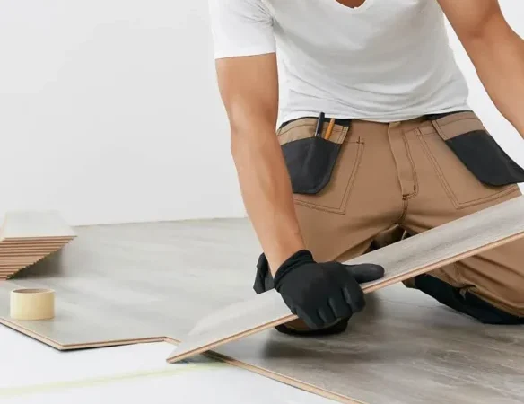 A person wearing safety glasses, gloves, and a tool belt installs laminate flooring, fitting a plank into place on a partially completed floor.