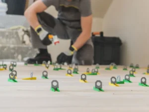 A worker installs floor tiles using a tile leveling system with green and yellow clips, kneeling in the background with blurred tools and a toolbox nearby.