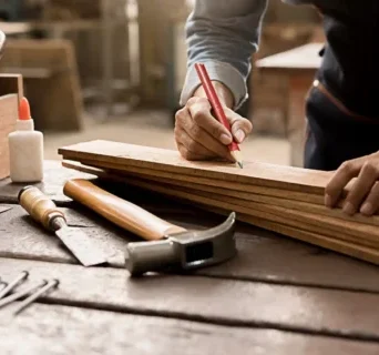 A person marks wooden planks with a pencil on a workbench, surrounded by woodworking tools—a scene showcasing skilled carpentry services with a hammer, chisel, glue, and nails at the ready.