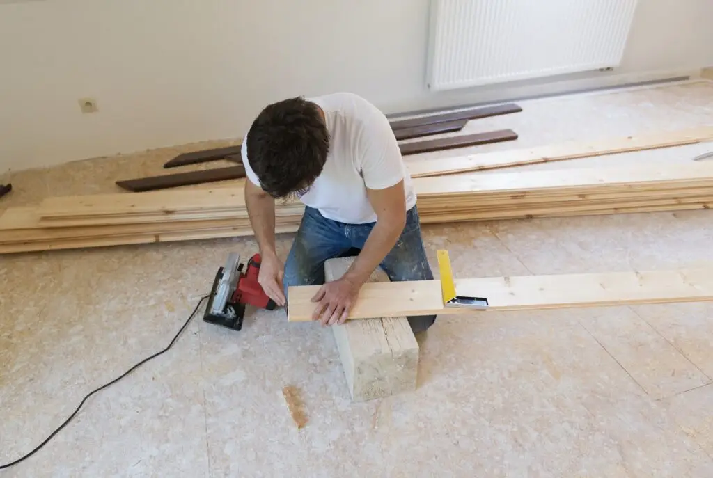 A person measures a wooden plank with a tape measure and a square tool while kneeling on the floor. Several wooden planks and a power tool are nearby. The room appears to be under renovation.