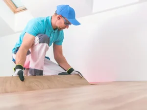 A worker wearing gloves, a blue cap, and safety glasses is kneeling on the floor, installing wooden laminate flooring in a bright, white room.
