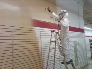 A person wearing protective gear stands on a ladder and spray paints a beige color onto a wall with horizontal slats in an indoor space, showcasing professional painting services.