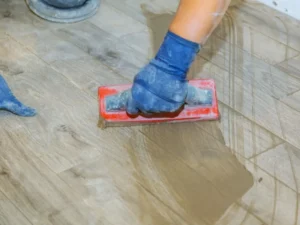 A person wearing a blue glove uses a red-handled float tool to spread grout over wood-look tiles during floor installation.