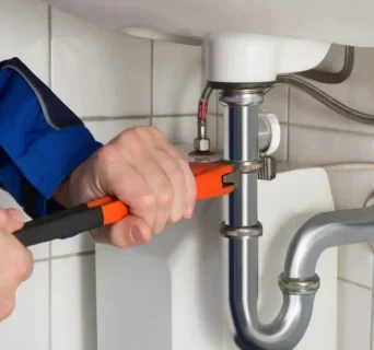 An expert handyman uses an orange pipe wrench to tighten or loosen a pipe under a white sink in a tiled bathroom.