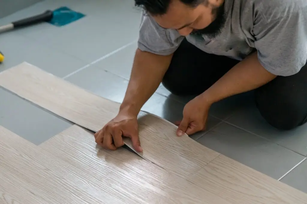 A handyman kneels on a tiled floor, carefully aligning and installing light wood-look vinyl planks, using their hands to press the flooring into place.