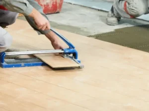 A construction worker in a red helmet cuts a ceramic tile with a manual tile cutter, showcasing professional tiling services, while another worker installs tiles in the background on a partially finished floor near large windows.