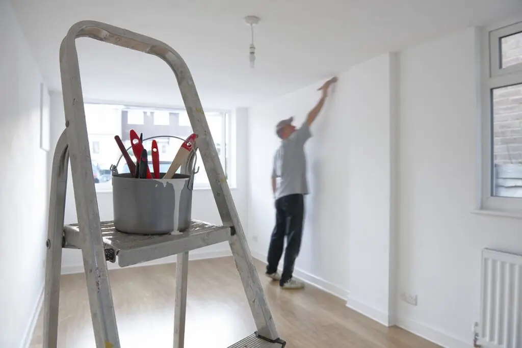 A bucket filled with painting tools sits on a stepladder in the foreground, while a person from professional painting services paints a white wall in a bright, unfurnished room with wooden floors.