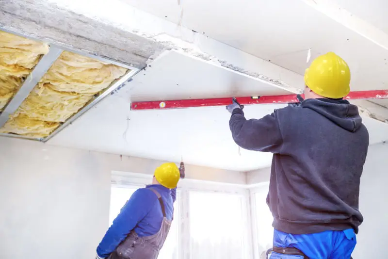 Two construction workers in hard hats install ceiling panels, using a spirit level to ensure alignment. Insulation is visible above, showcasing precise drywall services. The room is well-lit with natural light from large windows.