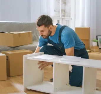 A man in blue overalls kneels on the floor and assembles white furniture in a living room surrounded by moving boxes.
