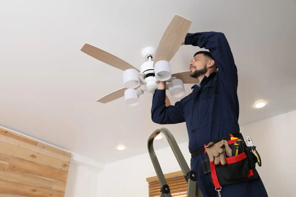A person wearing a tool belt and blue coveralls stands on a ladder, installing or repairing a ceiling fan with lights in a modern, well-lit room.