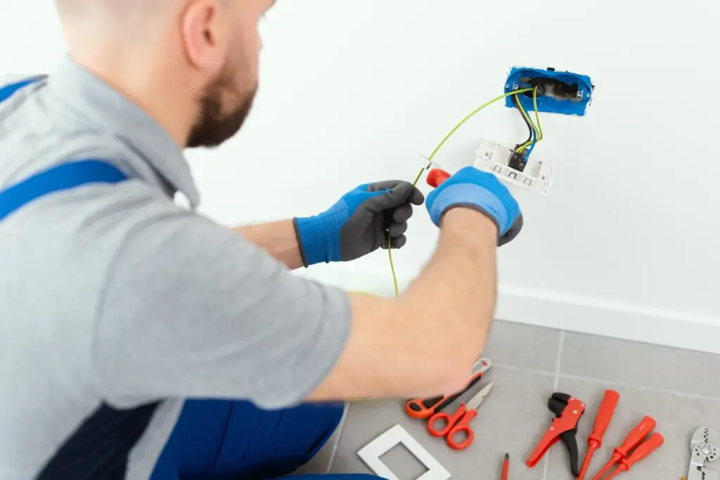An electrician wearing gloves works on wiring an electrical outlet in a wall, surrounded by various tools on a tiled floor, delivering expert electrical services.