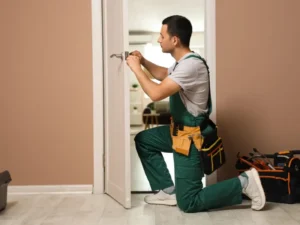 A handyman in green overalls kneels while fixing a door lock with tools, working attentively on home repairs inside, demonstrating general handyman services offered by BrightWise Handyman Services.