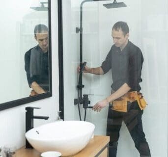 A plumber wearing a tool belt stands inside a glass shower, adjusting the shower controls during a bathroom remodeling. A white sink and black faucet are visible on the wooden countertop in the foreground.