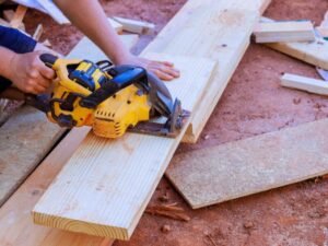 A carpenter uses a yellow circular saw to cut a wooden plank on a construction site, surrounded by sawdust and other pieces of wood, demonstrating the carpentry services offered by BrightWise Handyman.