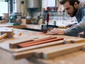 A man in a workshop uses a clamp and hand tool to work on a piece of wood, focusing intently. Various woodworking tools and wooden pieces are visible on the workbench and in the background, demonstrating the carpentry services offered by BrightWise Handyman.