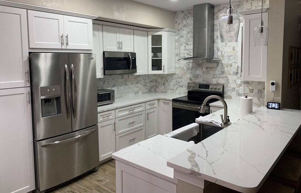 Modern kitchen with white cabinets, stainless steel appliances, marble countertops, and a marble backsplash. There are pendant lights above the breakfast bar and wooden flooring. The space appears clean and well-lit.
