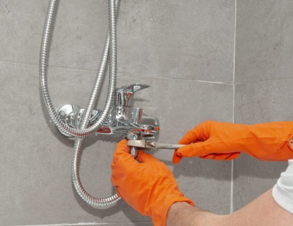 A person wearing orange gloves uses a wrench to tighten or repair a chrome shower faucet attached to a tiled bathroom wall.