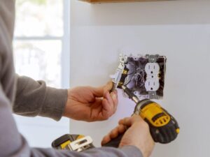 A person uses a power drill to perform a light switch repair next to an electrical outlet on a white wall. Exposed wires and electrical components are visible in the wall box.