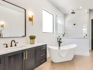 Modern bathroom with a black vanity, double sink, gold fixtures, a freestanding white tub with a towel, a glass-enclosed shower, large mirror, wall sconces, and a potted plant on the counter.