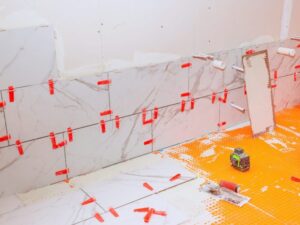White marble tiles being installed on a wall with orange underlayment on the floor. Red tile spacers and clips are visible between tiles. A white bucket and tile-cutting tools are nearby.