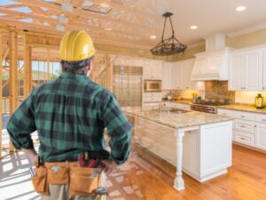 A construction worker with a tool belt and hard hat looks at a partially built wooden structure that transitions into a finished, modern white kitchen with granite countertops and wood floors.